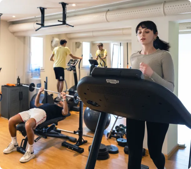 Persone che si allenano in palestra con tapis roulant, pesi e attrezzi fitness.