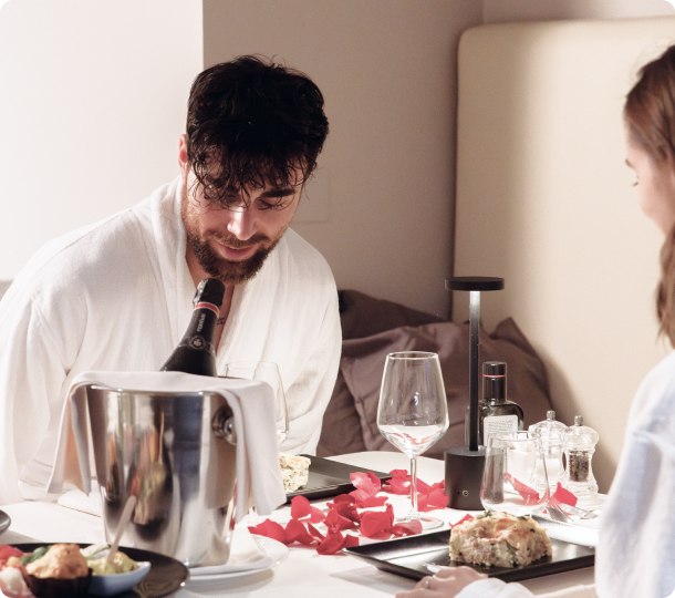 Una coppia in accappatoio fa colazione romantica a letto con vino e petali di rosa.